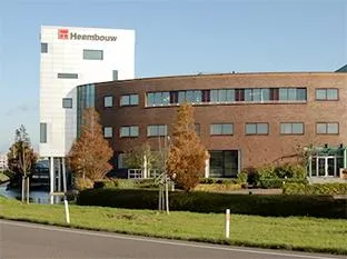 En kontorbygning i røde mursten med et hvidt tårn, der bærer Heembouw-logoet, beliggende ved en vej og et grønt område.